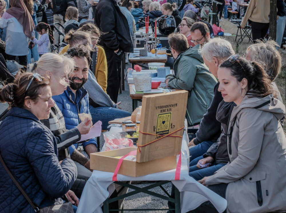 Viele Menschen sitzen auf Biertischgarnituren im Freien an einem schönen Tag. Manche sind ins Gespräch vertieft, manche stehen auch.