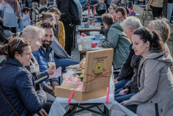 Viele Menschen sitzen auf Biertischgarnituren im Freien an einem schönen Tag. Manche sind ins Gespräch vertieft, manche stehen auch.