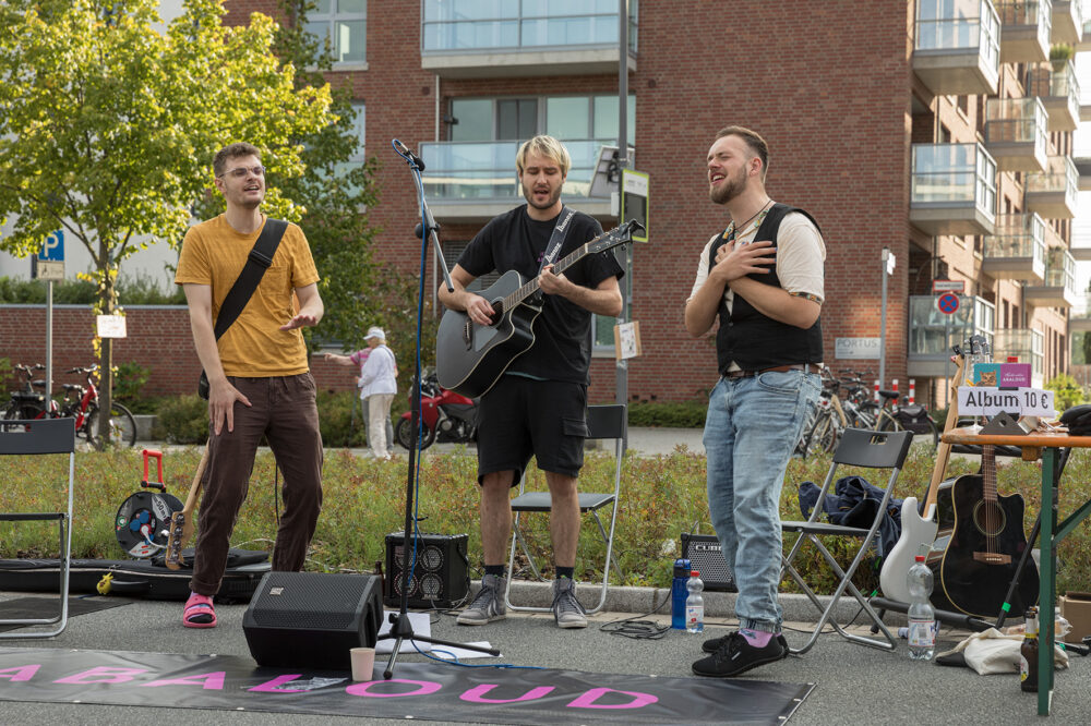 Drei Männer musizieren. Sie stehen auf der Straße. Zwei singen und einer spielt Gitarre. 