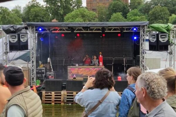 Publikum steht vor einer großen schwarzen Bühne. Dahinter ist ein Fluss, der übers Ufer gegangen ist und die Bühne fast umspült