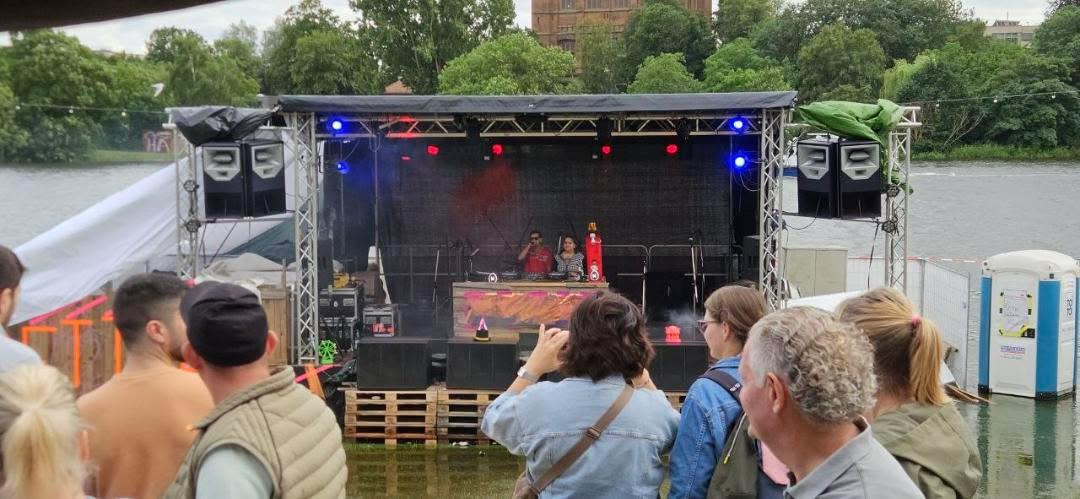 Publikum steht vor einer großen schwarzen Bühne. Dahinter ist ein Fluss, der übers Ufer gegangen ist und die Bühne fast umspült