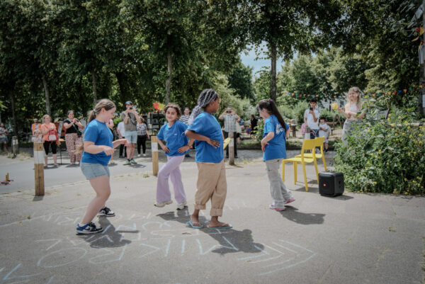 Auf einer Außenfläche sind Markierungen mit Kreide aufgemalt. Auf ihnen tanzen vier Jugendliche in blauen T-Shirts. Im hintergrund sind weitere Personen zu sehen und große Bäume mit grünem Laub.