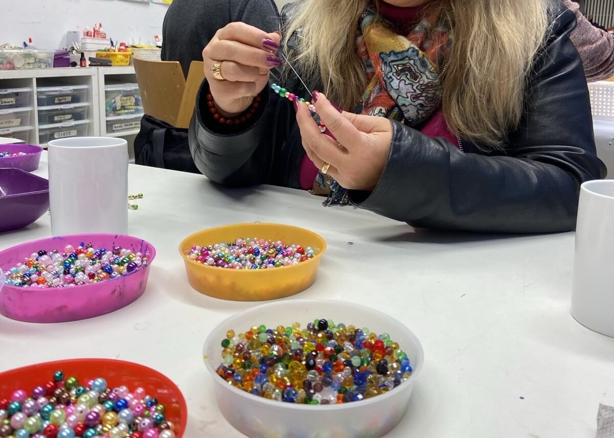 Bunte Plastikperlen liegen in Schalen auf einem Tisch, es sind zwei Hände zusehen, die damit basteln.