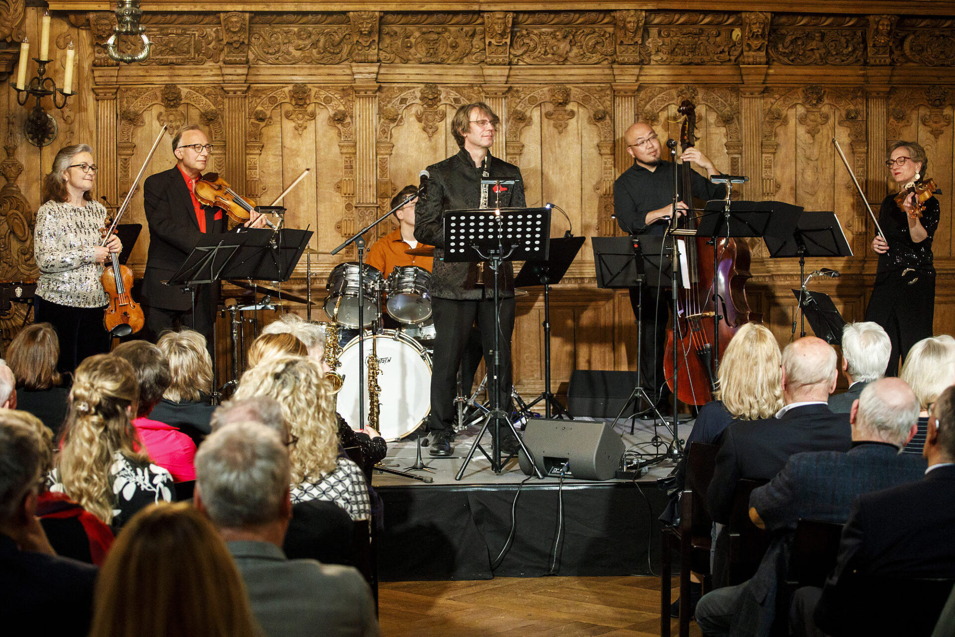 Publikum sitzt in Stuhlreihen mit Mittelgang und ist von Hinten zu sehen. Auf einer niedrigen Bühne befinden sich sechs Musiker:innen mit klassischen Instrumenten. Die Wand hinter hnen ist holzverziert.