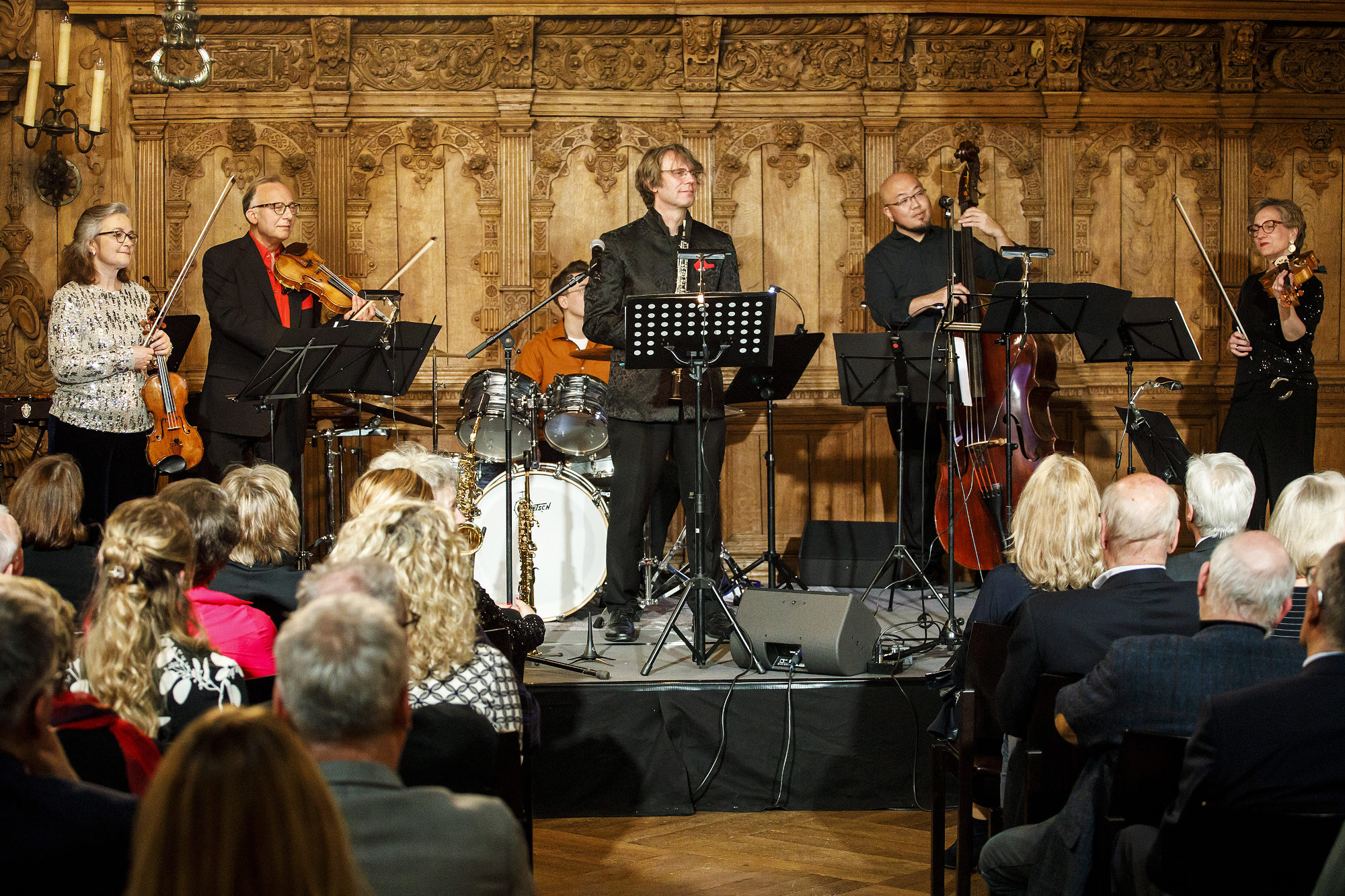 Publikum sitzt in Stuhlreihen mit Mittelgang und ist von Hinten zu sehen. Auf einer niedrigen Bühne befinden sich sechs Musiker:innen mit klassischen Instrumenten. Die Wand hinter hnen ist holzverziert.