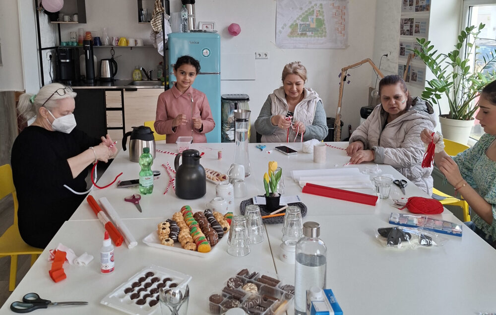 Auf einem weißen Tischblock befinden sich Süßigkeiten, Thermoskannen und buntes Papier. Rund um die Tische sitzen mehrere Personen, die am Basteln sind.