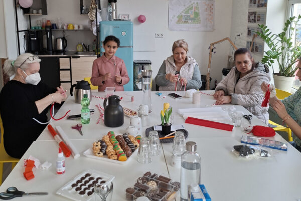 Auf einem weißen Tischblock befinden sich Süßigkeiten, Thermoskannen und buntes Papier. Rund um die Tische sitzen mehrere Personen, die am Basteln sind.