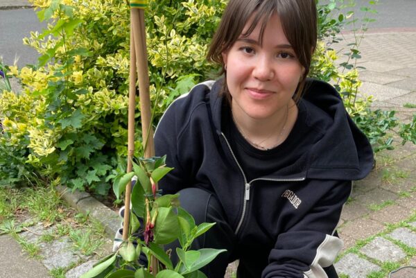 Eine Person hockt auf dem Boden. Sie schaut in die Kamera. Mit einer Hand hält sie eine Pflanze, die sie in einen Eimer einpflanzt.