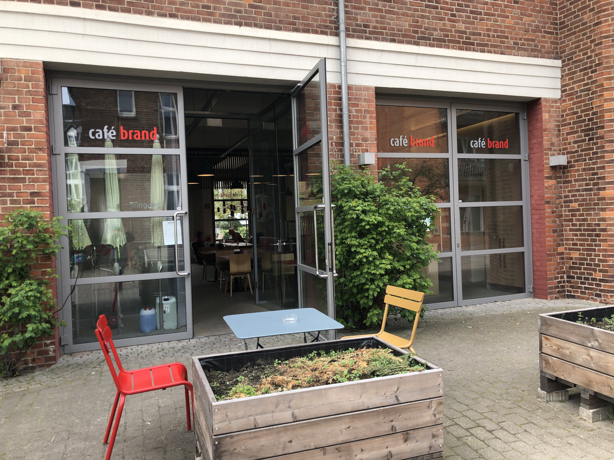 Vor großen Fenstern und einer Glastür befindet sich ein kleiner Tisch mit zwei Stühlen. Davor ist ein nichtbepflanztes Hochbeet.