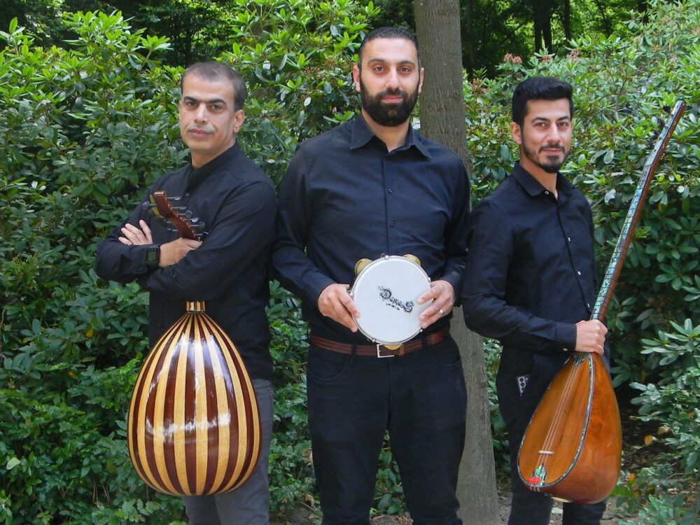 Drei Personen stehen nebeneinander im Grünen. Sie tragenschwarze Hemden und halten traditionelle Musikinstrumente in der Hand.