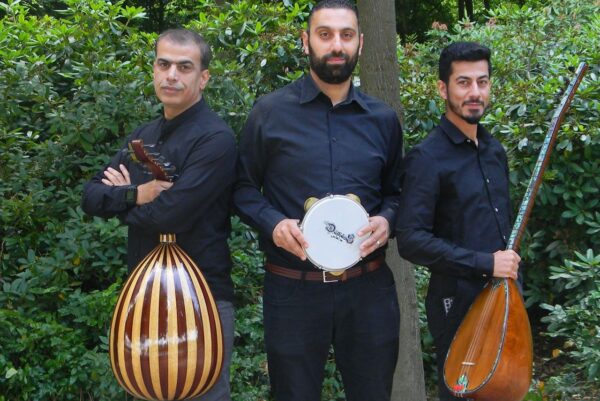 Drei Personen stehen nebeneinander im Grünen. Sie tragenschwarze Hemden und halten traditionelle Musikinstrumente in der Hand.