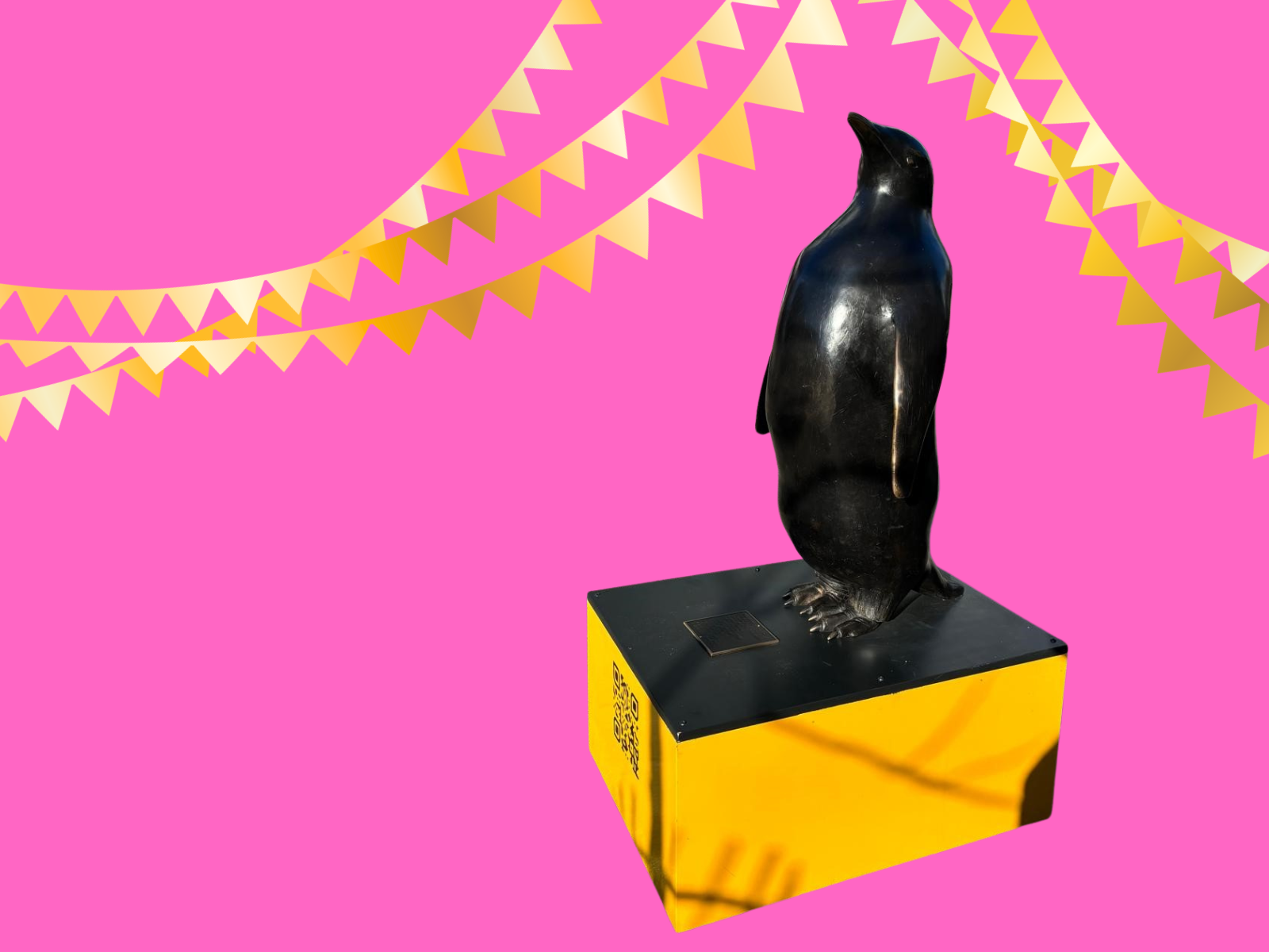 Vor einem Pinken Hintergrund ist eine Bronzeskulptur in Form eines Pinguins auf einem Sockel zu sehen. Oben im Bild sind Wimpelketten