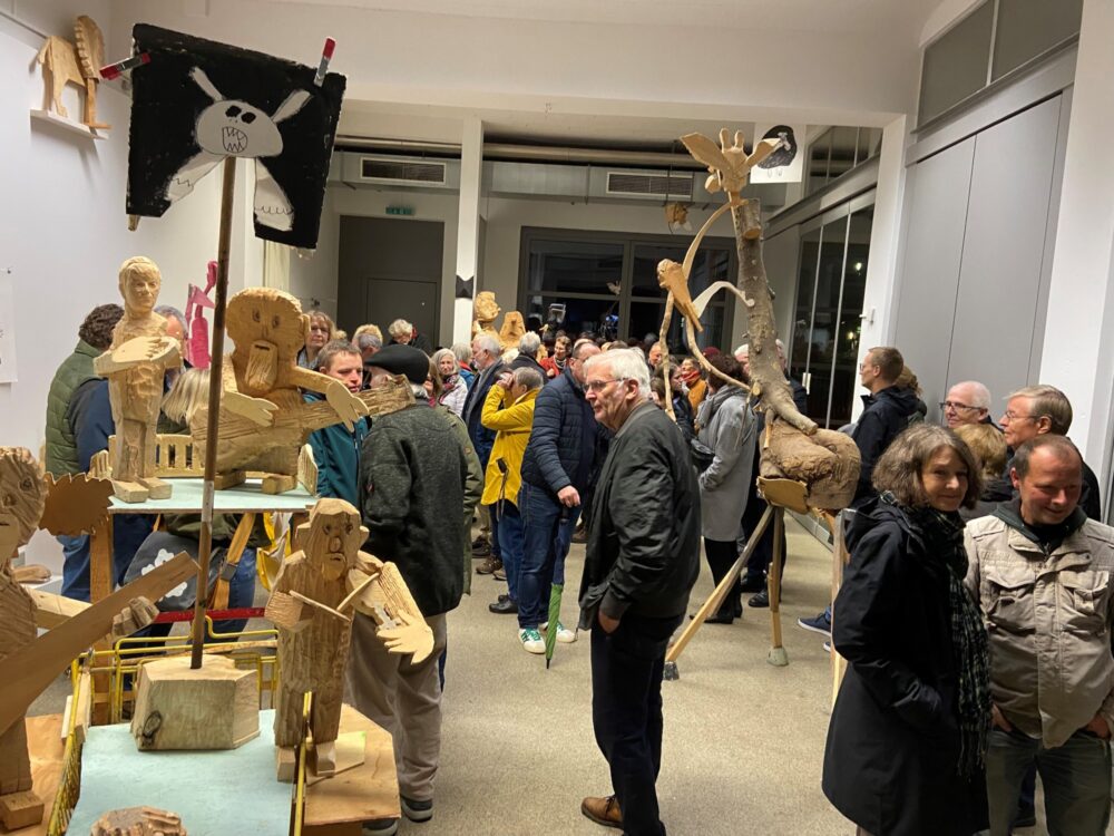 Bei einer Vernissage sind in einer Galerie viele Skulpturen aus Holz zu sehen, die teilweise auf dem Boden und teilweise auf Sockeln zu sehen sind. Zwischen den Skulpturen befinden sich viele Menschen, die zur Eröffnung der Ausstellung gekommen sind.