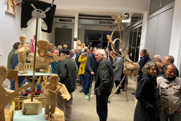 Bei einer Vernissage sind in einer Galerie viele Skulpturen aus Holz zu sehen, die teilweise auf dem Boden und teilweise auf Sockeln zu sehen sind. Zwischen den Skulpturen befinden sich viele Menschen, die zur Eröffnung der Ausstellung gekommen sind.