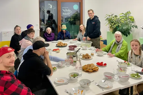 Rund um einen großen weißen Tischblock sitzen viele Personen. Vor ihren befindet sich Geschirr und Essen. Eine Person steht.