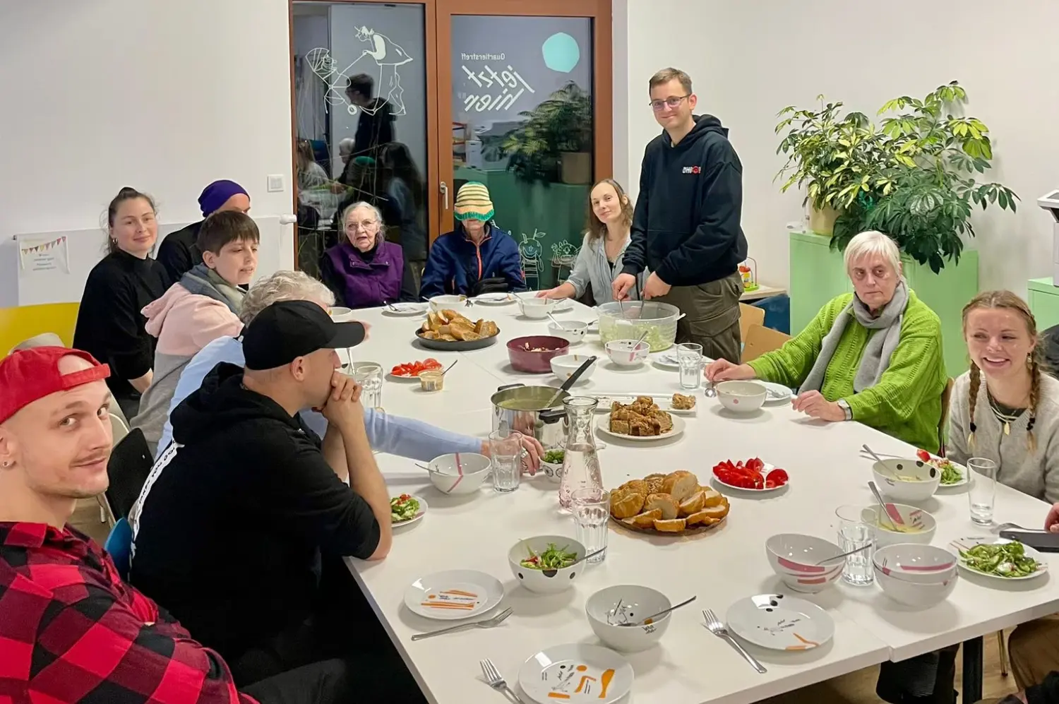 Rund um einen großen weißen Tischblock sitzen viele Personen. Vor ihren befindet sich Geschirr und Essen. Eine Person steht.