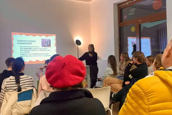 Menschen sitzen in Stuhlreihen und sind von hinten zu sehen. Sie Blicken auf eine Leinwandpräsentation. Eine Person steht daneben und spricht.