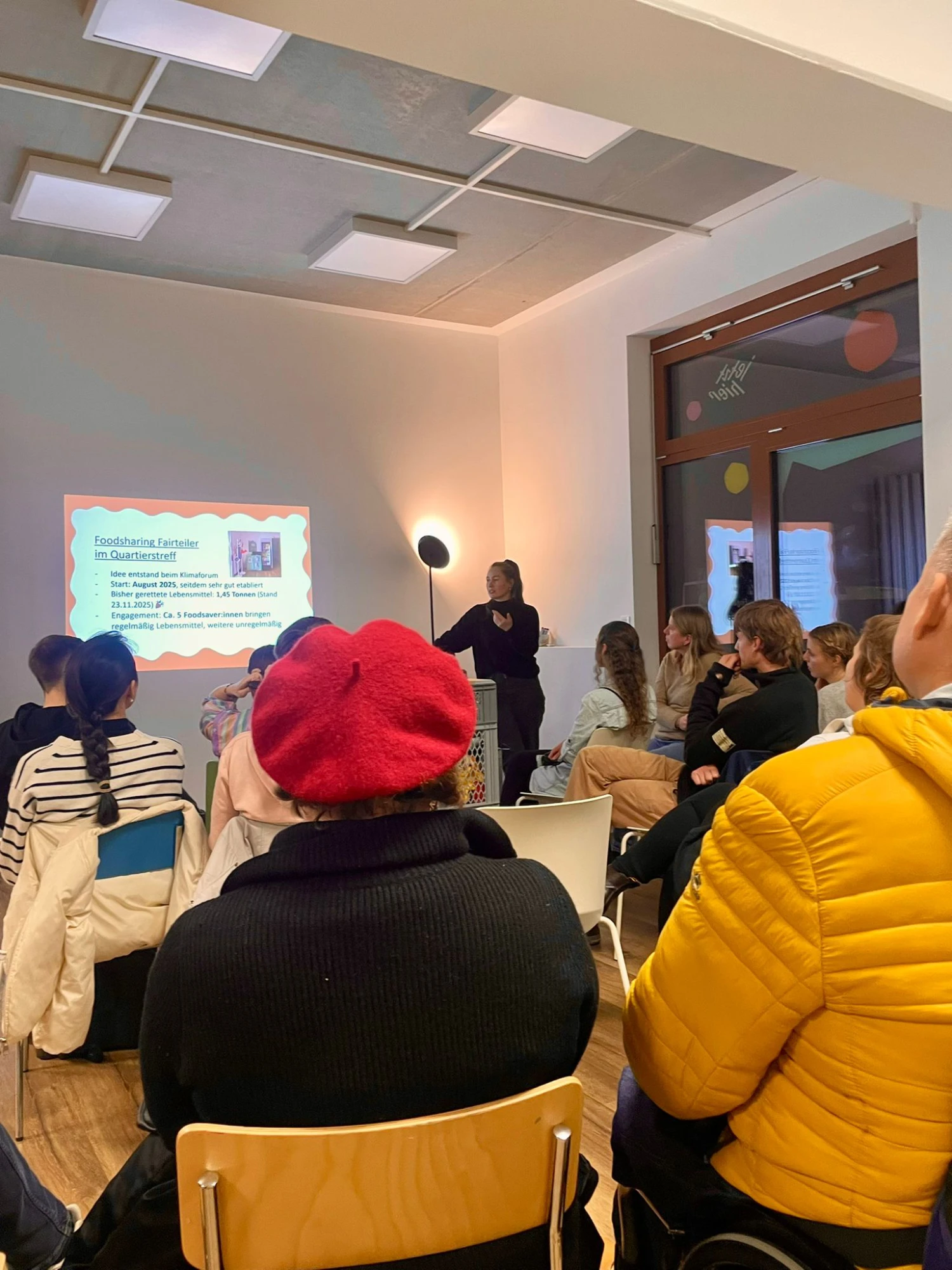 Menschen sitzen in Stuhlreihen und sind von hinten zu sehen. Sie Blicken auf eine Leinwandpräsentation. Eine Person steht daneben und spricht.