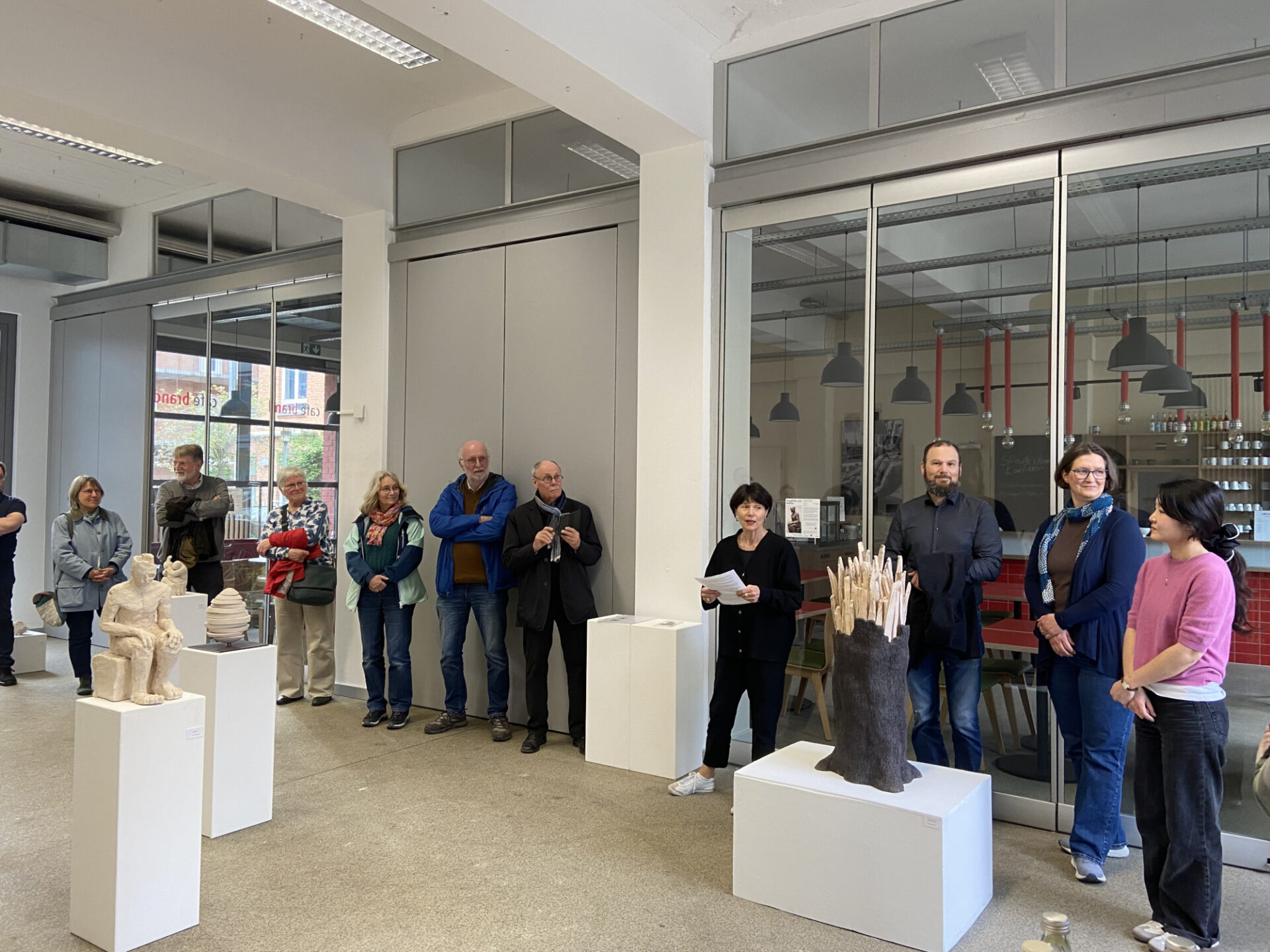 In einer Galerie sind Kunstobjekte aus Ton auf weißen Sockeln zu sehen. Eine Person hält Papier in der Hand und spricht. Rundherum stehen weitere Personen im Raum und hören zu.