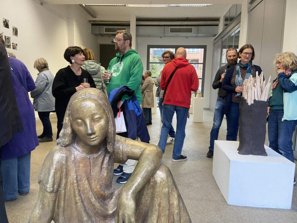 In einer Galerie befinden sich Menschen, verteilt in Grüppchen zwischen Kunstobjekten ins Gespräch vertieft. Ganz vorne ist eine Bronzefigur im Bild.