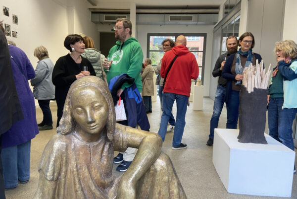 In einer Galerie befinden sich Menschen, verteilt in Grüppchen zwischen Kunstobjekten ins Gespräch vertieft. Ganz vorne ist eine Bronzefigur im Bild.
