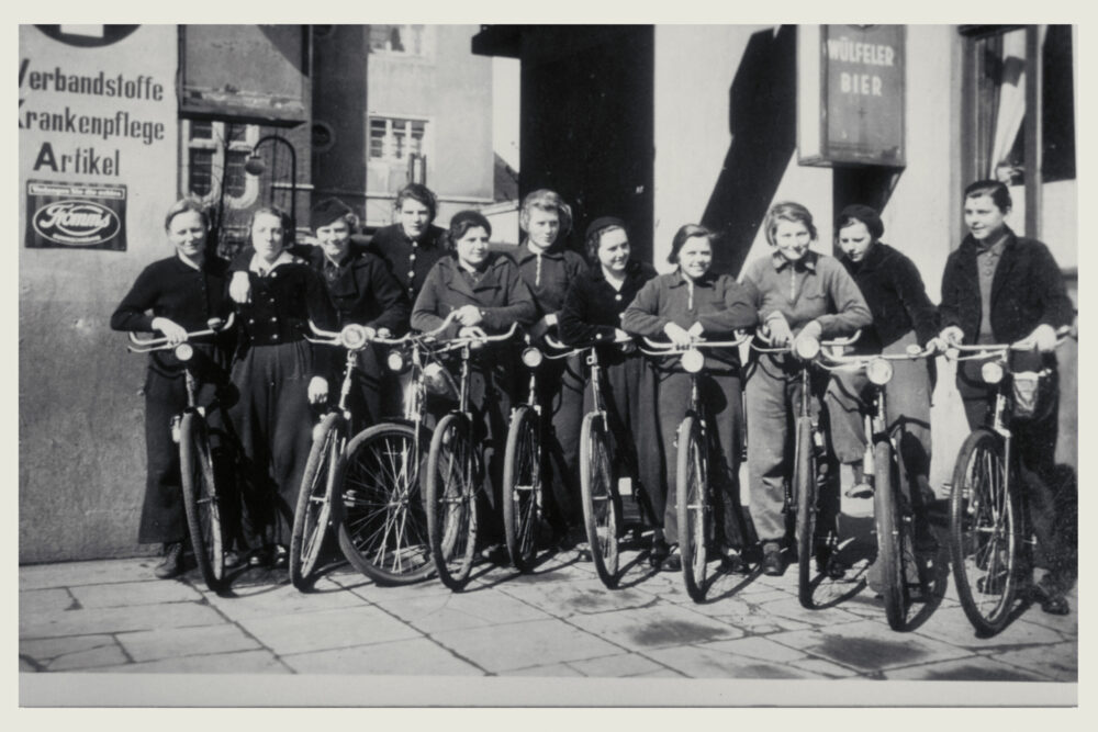 Dies ist eine historische Aufnahme von Fahrradfahrerinnen mit ihren Rädern in einer Toreinfahrt. Das Foto ist schwarz weiß