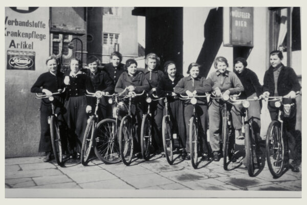 Dies ist eine historische Aufnahme von Fahrradfahrerinnen mit ihren Rädern in einer Toreinfahrt. Das Foto ist schwarz weiß