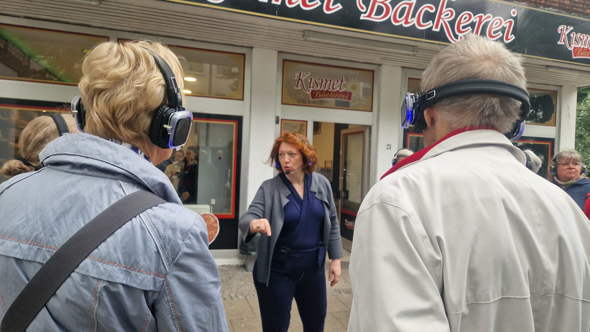 Eine Person mit Kopfhörern und Headset gestikuliert vor mehreren Personen, uns mit dem Rücken zugewandt, die ebenfalls Kopfhörer tragen. Dahinter befindet sich eine Bäckerei, die Kismet heißt.