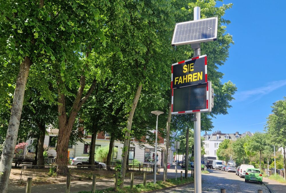 Entlang einer Straße säumen links große Laubbäume den gepflasterten Weg. Weiter hinten sind parkende Autos zu sehen. Vorne befindet sich eine Geschwindigkeitsmessung. Auf einem Schild steht "Sie fahren" Darunter befindet sich die Tempoanzeige.