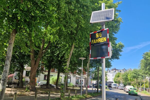 Entlang einer Straße säumen links große Laubbäume den gepflasterten Weg. Weiter hinten sind parkende Autos zu sehen. Vorne befindet sich eine Geschwindigkeitsmessung. Auf einem Schild steht "Sie fahren" Darunter befindet sich die Tempoanzeige.
