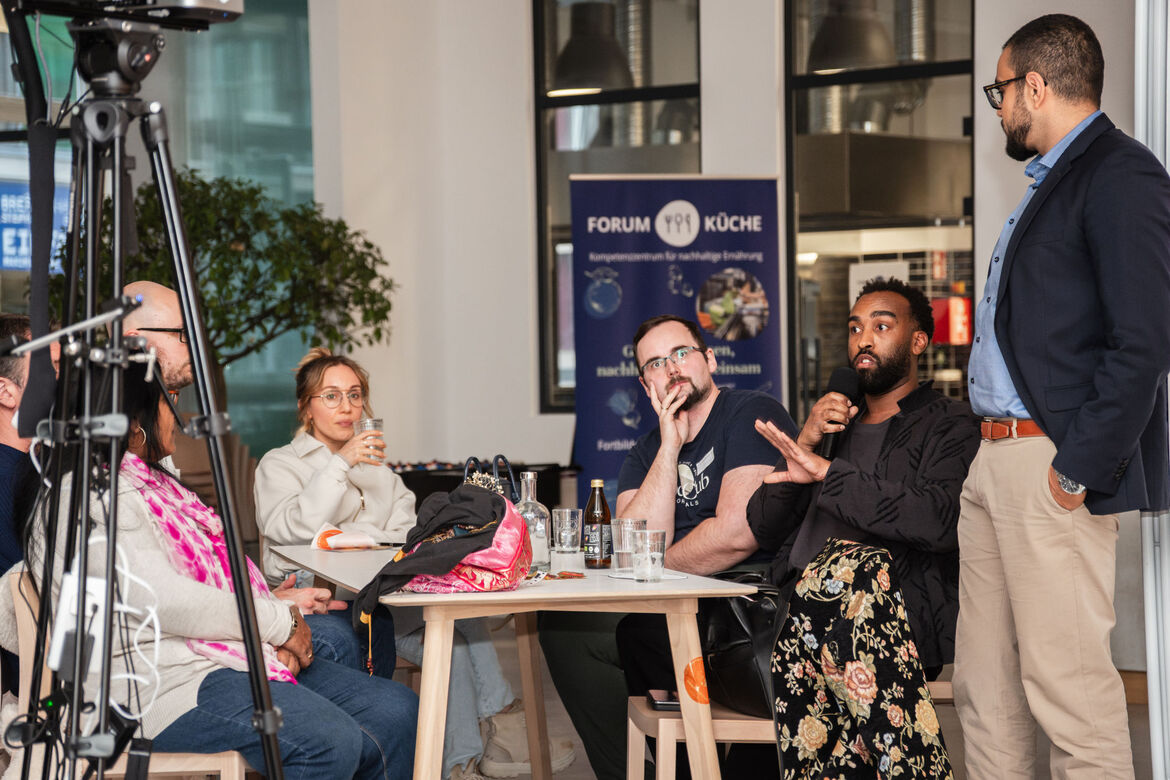 Mehrere Personen sitzen rund um einem kleinen Tisch, eine weitere Person steht rechts von Ihnen. Eine Person spricht.