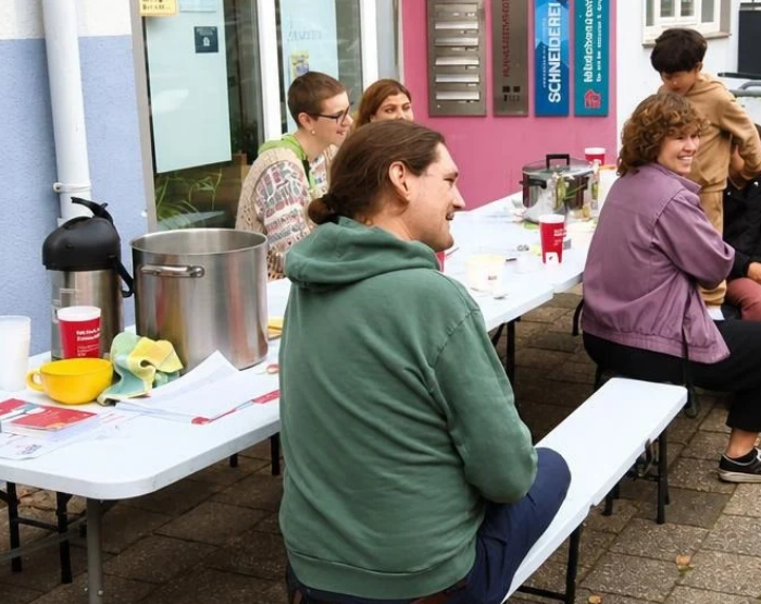 An einer Biertischgarnitur mit Topf und Kaffeekanne sitzen lächende Menschen.