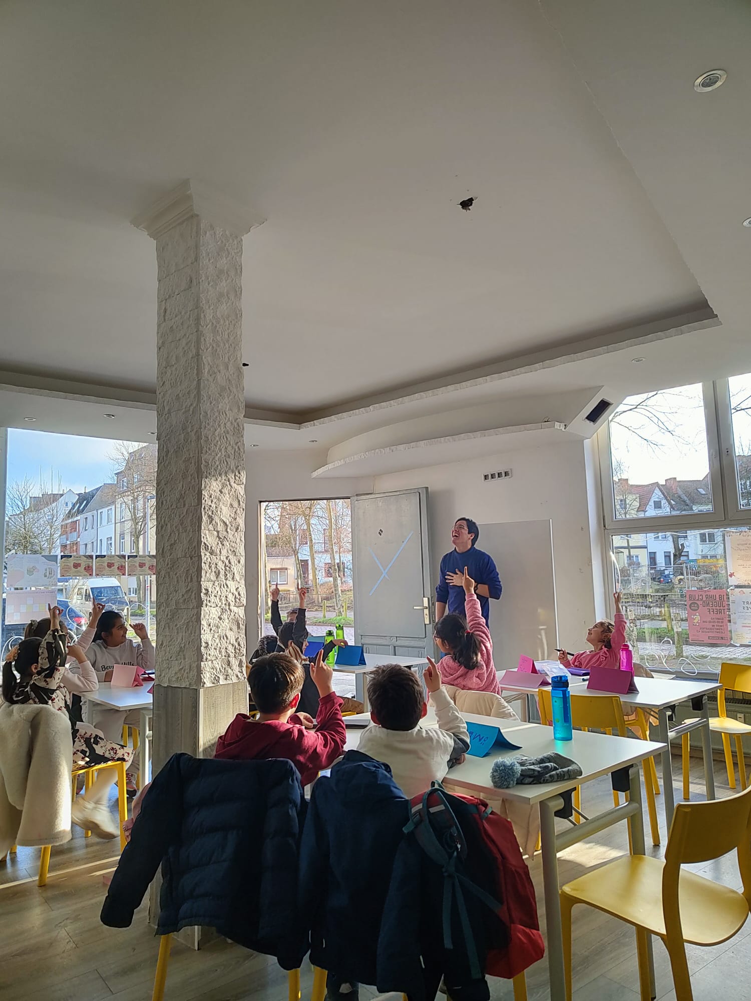 In einemNachbarschaftstreff sitzen Kinder an Tischen auf Stühlen und schauen einer lachenden Person zu.