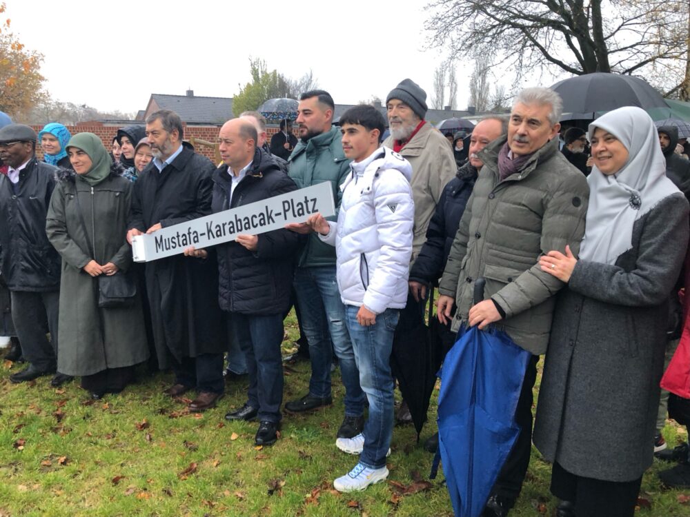 Menschen halten das Straßenschild "Mustafa-Karabacak-Platz" bei der einweihung des Platzes.