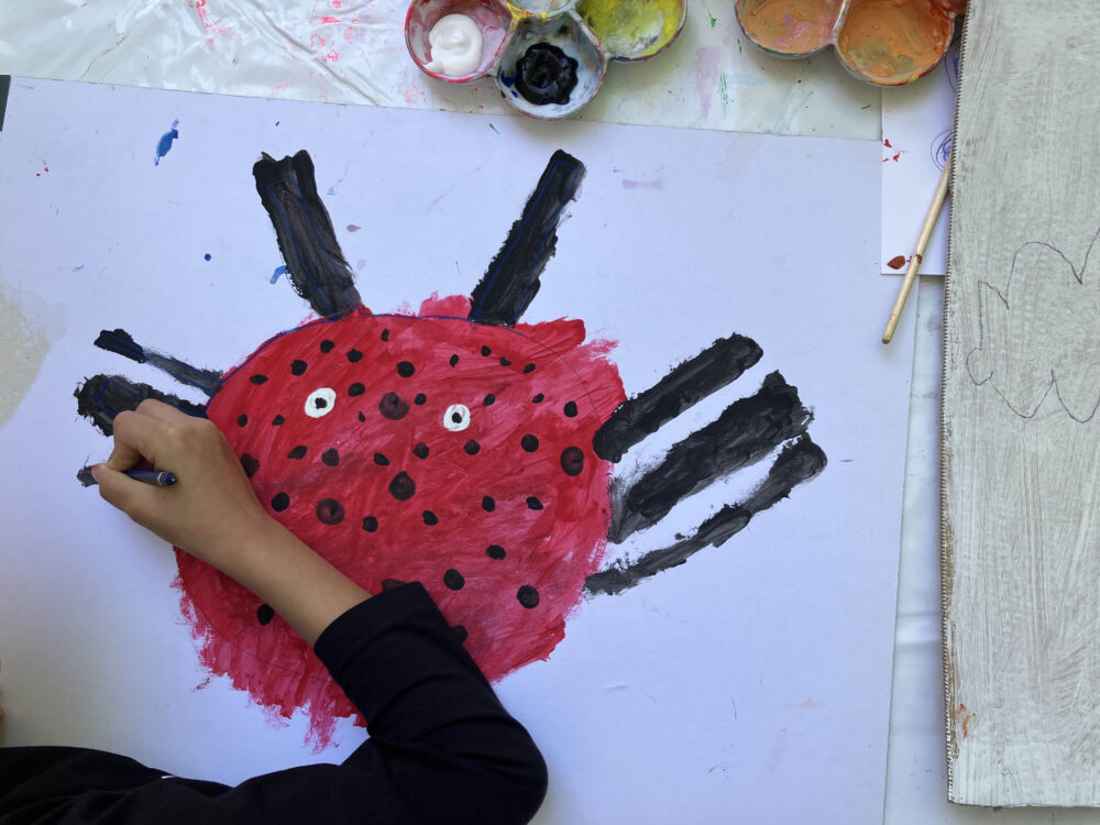 Eine Ansicht von Oben auf einen Tisch mit großem Blatt Papier, auf das eine Hand Farbe aufträgt. Dargestellt wird ein Käfer. Oberhalb des Blattes sind Farbbecher zu sehen