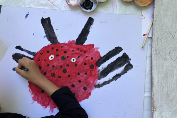 Eine Ansicht von Oben auf einen Tisch mit großem Blatt Papier, auf das eine Hand Farbe aufträgt. Dargestellt wird ein Käfer. Oberhalb des Blattes sind Farbbecher zu sehen