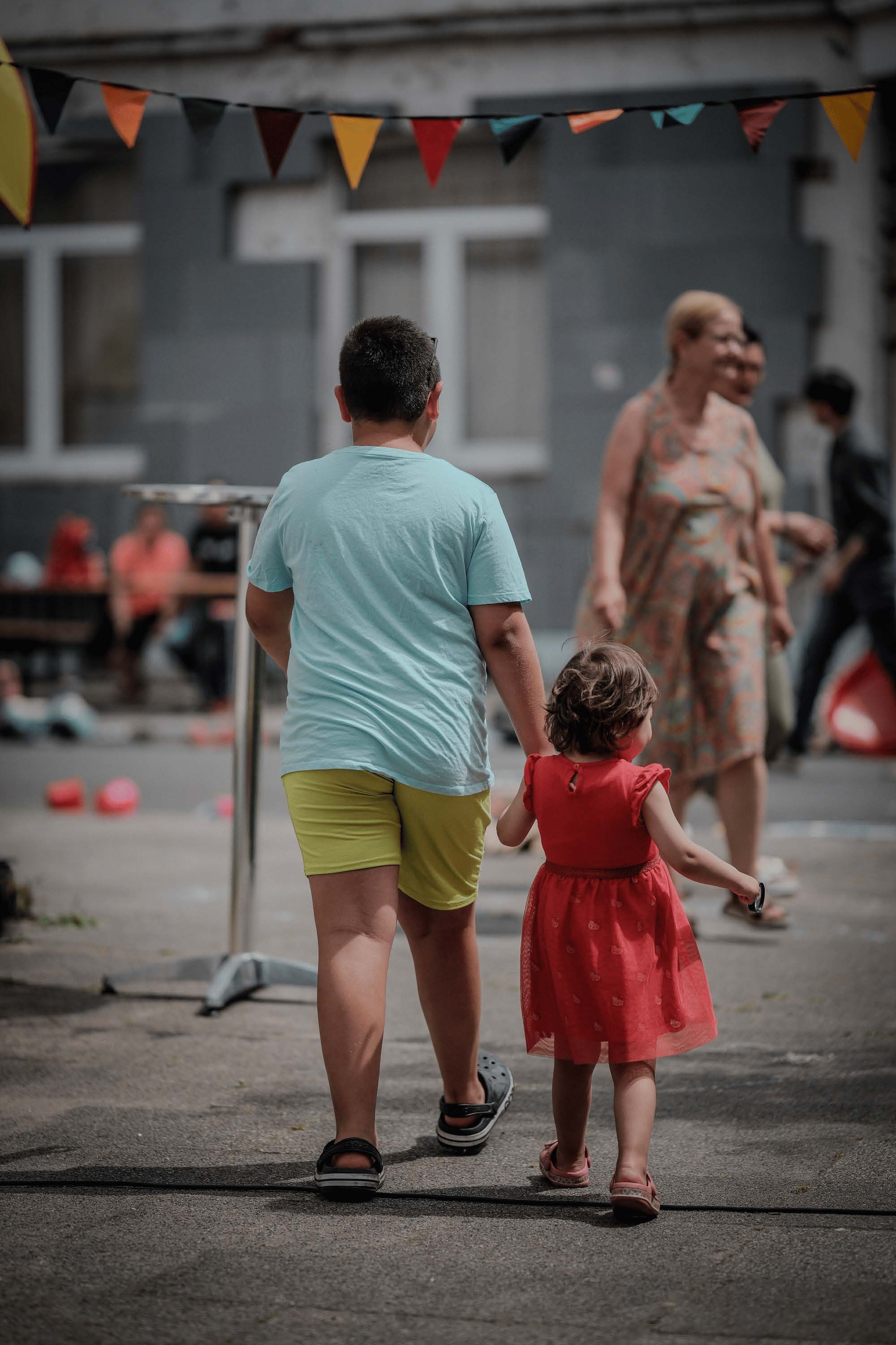 Ein Junge in hellblauem T-Shirt und gelben Shorts geht Hand in Hand mit einem kleinen Mädchen im roten Kleid. Sie laufen im Freien an bunten Wimpeln und anderen Menschen vorbei.
