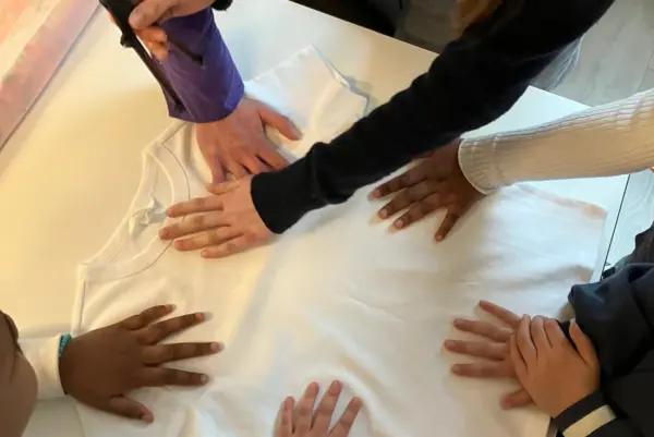 Ein weißes T-Shirt liegt auf einem Tisch. Es sind die Arme und Hände von mehreren Kindern zu sehen, die auf dem T-Shirt aufgelegt sind. In einer Hand ist ein Stift zu sehen.