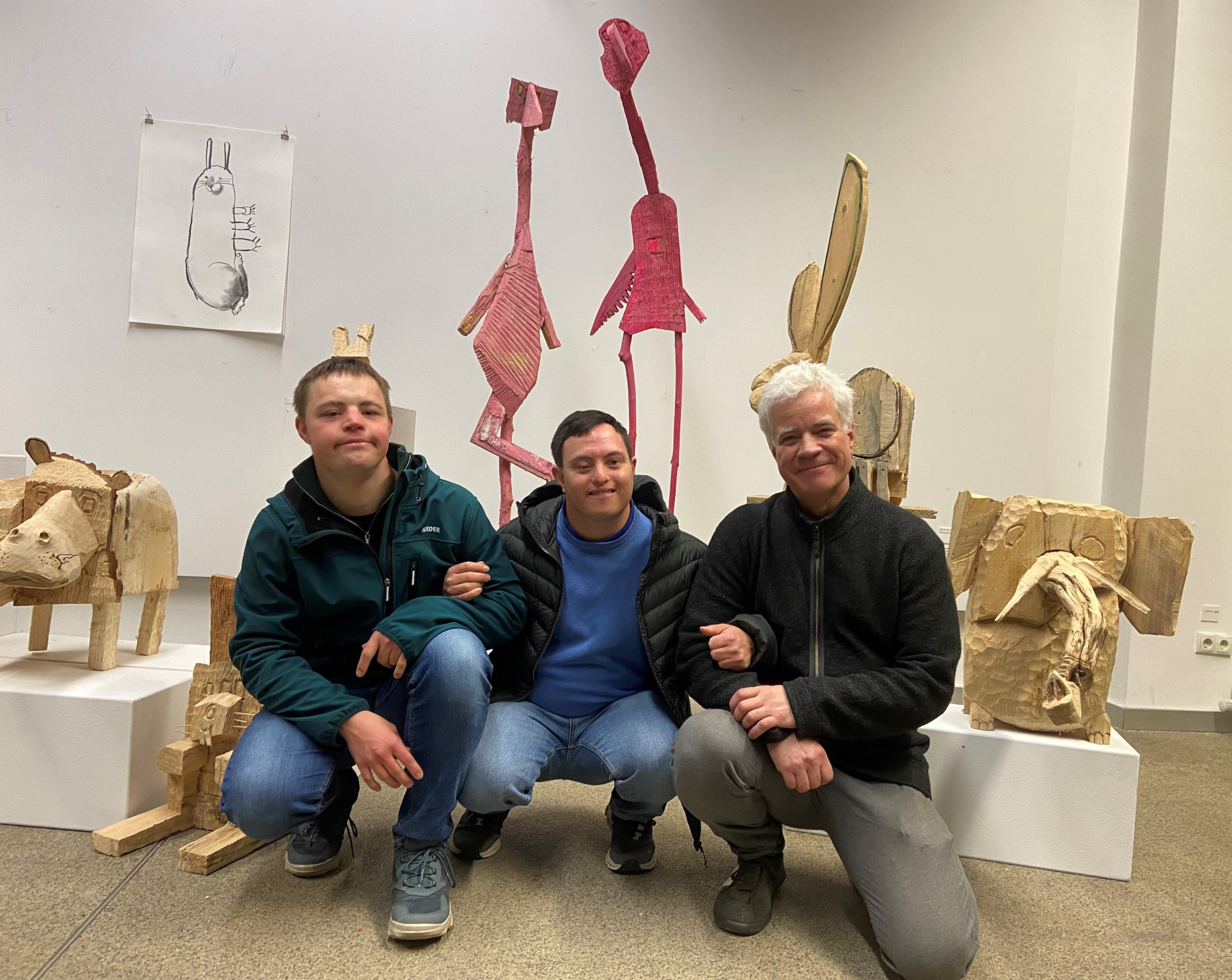 Die drei Künstler einer Ausstellung mit Skulpturen sind auf einem Gruppenfoto zwischen ihren Werken zu sehen. Für das Gruppenfoto haben sie sich auf den Boden gehockt und sich gegenseitig an den Armen untergehakt.