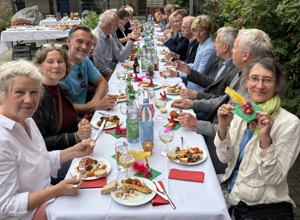 an einer langen festlich geschmückten Tatel draußen mit weißen Tischdecken sitzen Menschen und essen, reden oder lächeln