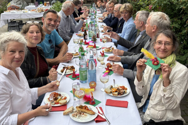 an einer langen festlich geschmückten Tatel draußen mit weißen Tischdecken sitzen Menschen und essen, reden oder lächeln