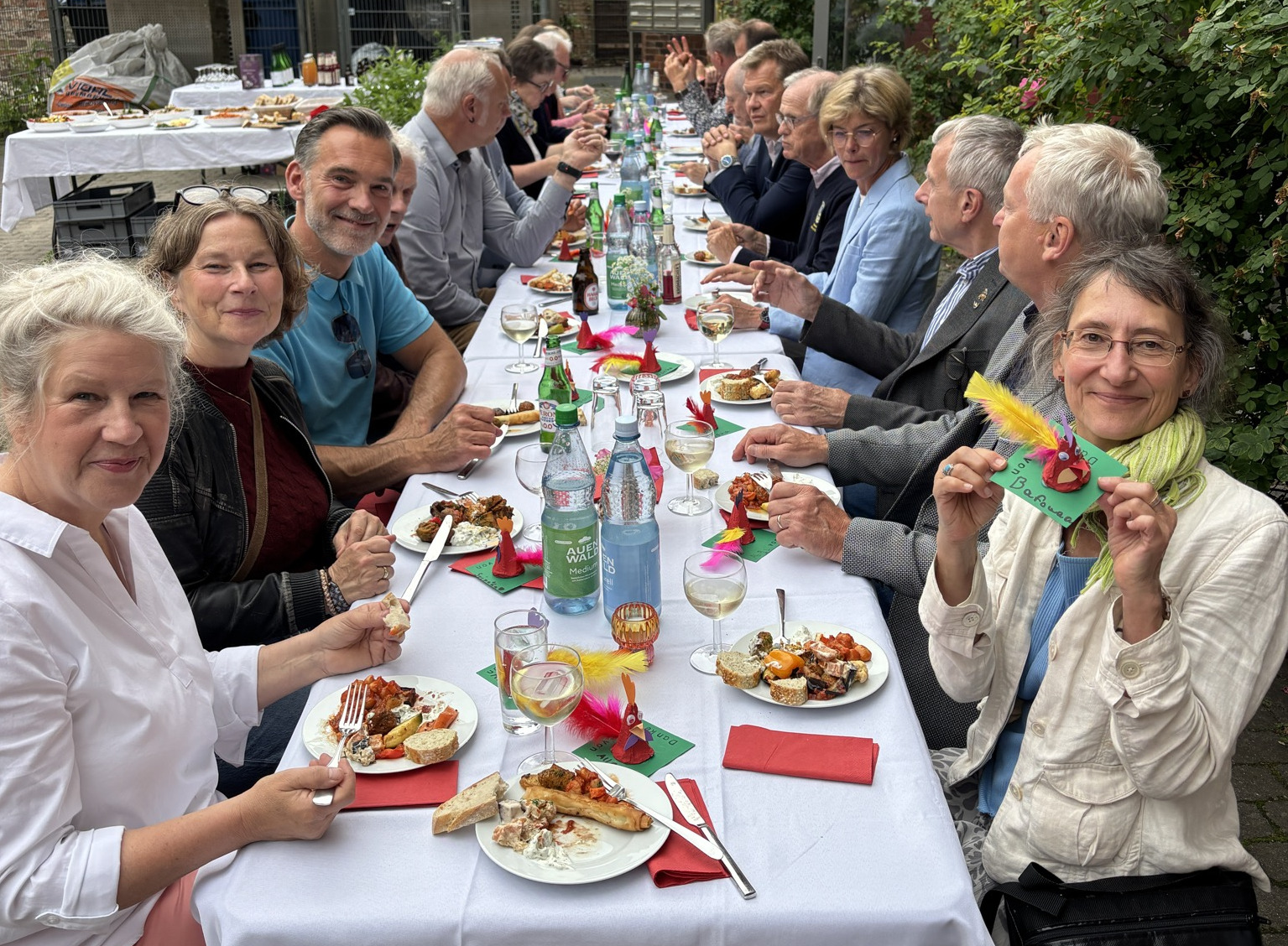 an einer langen festlich geschmückten Tatel draußen mit weißen Tischdecken sitzen Menschen und essen, reden oder lächeln