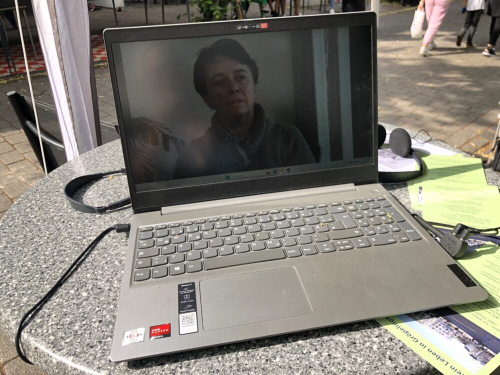Auf einem runden Bistrotisch befindet sich ein Laptop auf dem ein Video läuft. Unter und neben dem Laptop sind Flyer, dahinter liegt ein Kopfhörer. Auf dem Video ist eine Frau zu erkennen.