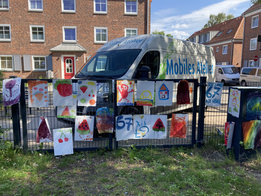 An einem Zaun hängen A3 Papiere, die mit Farben bemalt wurden. Dahinter ist ein Bulli zu sehen, an dem der Schriftzug "Mobiles Atelier" steht