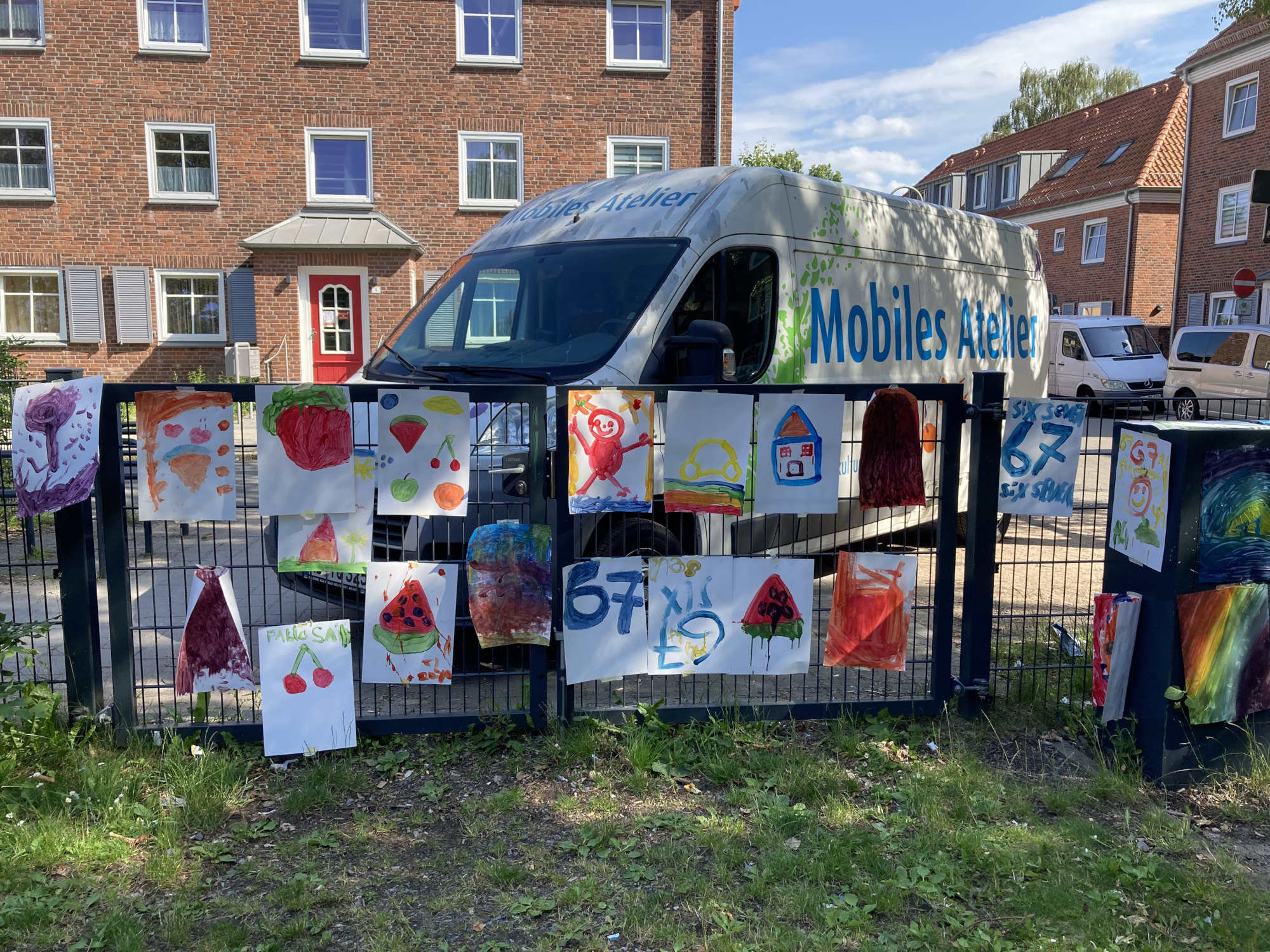 An einem Zaun hängen A3 Papiere, die mit Farben bemalt wurden. Dahinter ist ein Bulli zu sehen, an dem der Schriftzug "Mobiles Atelier" steht