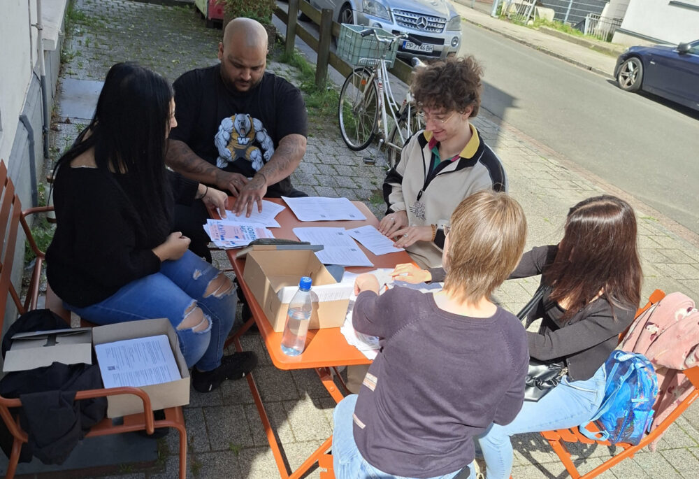 Fünf Personen sitzen vor einem Haus rund um einen Tisch. Sie sind ganz konzentriert dabei, Flugblätter zu falten.