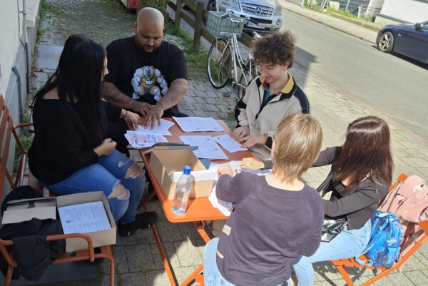 Fünf Personen sitzen vor einem Haus rund um einen Tisch. Sie sind ganz konzentriert dabei, Flugblätter zu falten.