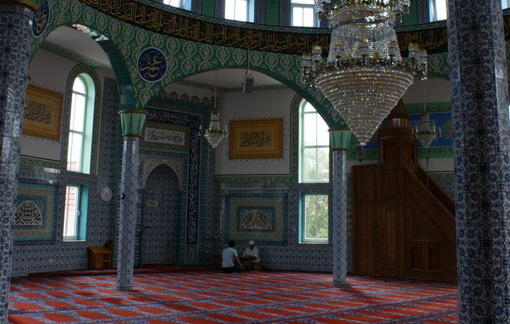 Der kunstvoll verzierte Gebetsraum der Fatih moschee ist zu sehen. Zwei Personen sitzen im Hintergrund.