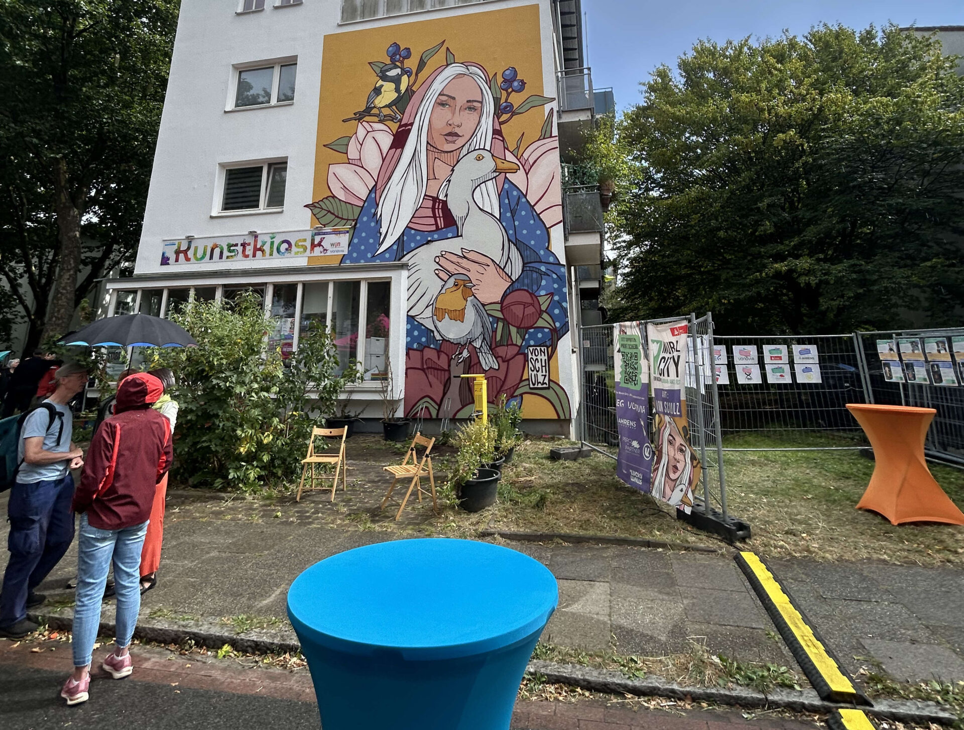 Eine mit einem Fassadengemälde verzierte Hausfassade ist zu sehen. Darauf hält eine Frauengestalt eine Gans im Arm. Vor dem Gebäude befinden sich links ein paar Personen.