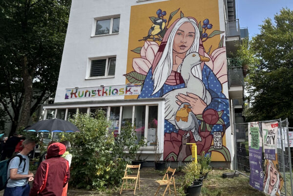 Eine mit einem Fassadengemälde verzierte Hausfassade ist zu sehen. Darauf hält eine Frauengestalt eine Gans im Arm. Vor dem Gebäude befinden sich links ein paar Personen.